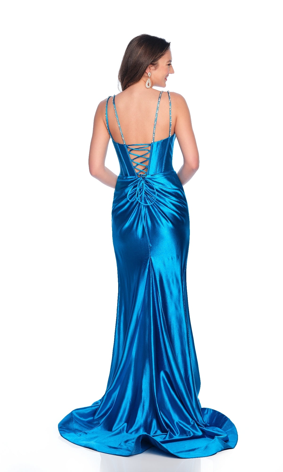 A woman faces away from the camera in the Peacock Blue Prom Dress: Dave & Johnny 12335, which features a lace-up corset bodice, thin straps, a shiny satin finish, and a high-leg front slit with the gown elegantly pooling on the floor.